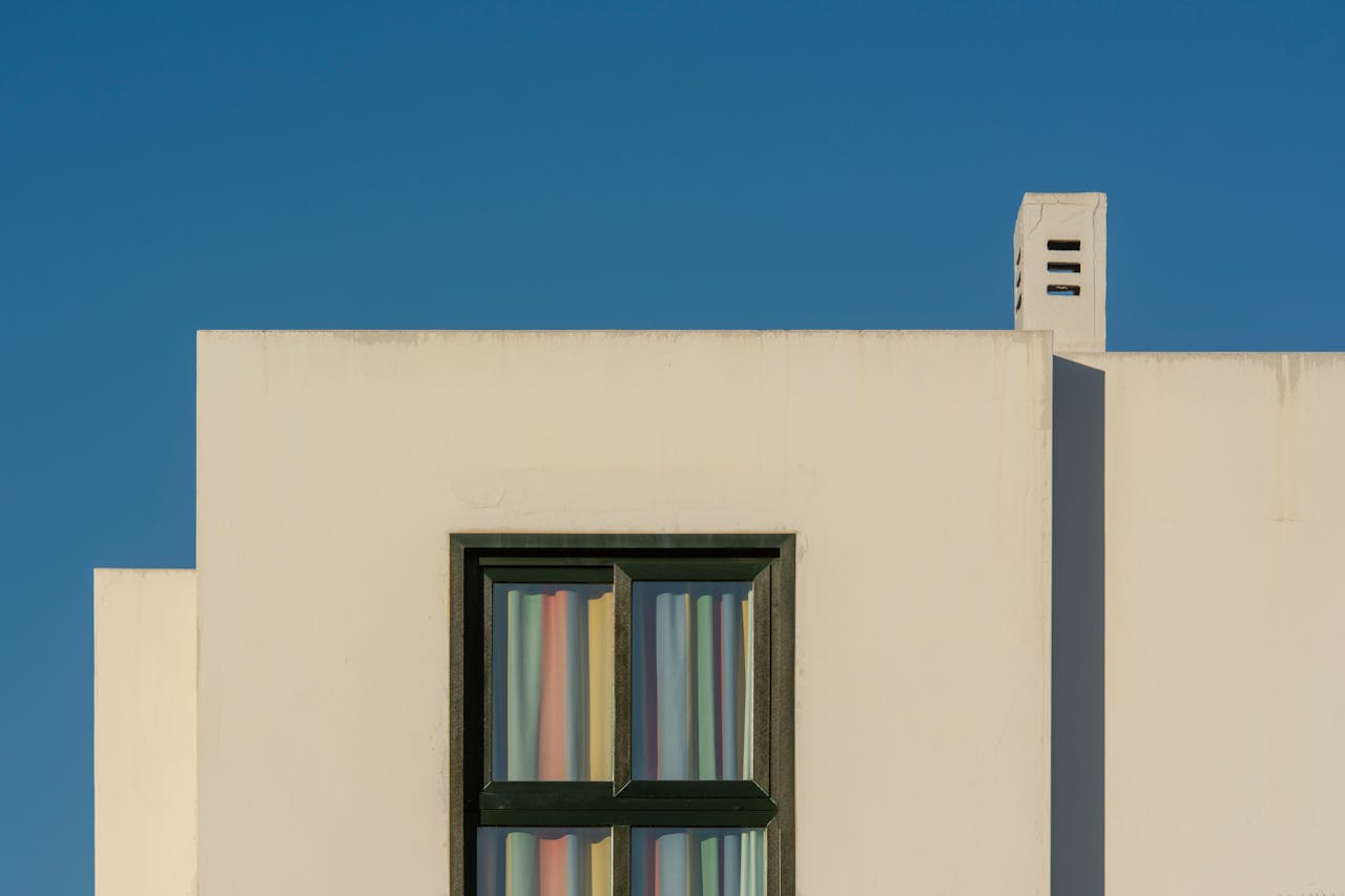 Minimalist facade of a modern building featuring a window with colorful curtains and a chimney against a blue sky.